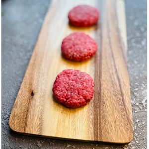 Three raw hamburger patties on a wooden cutting board with a gray background