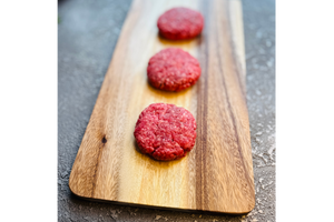 Three raw hamburger patties on a wooden cutting board with a gray background