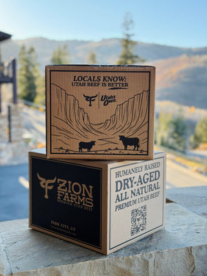 Two cardboard boxes from Zion Farms with mountain and cow graphics on a stone surface with a scenic background.
