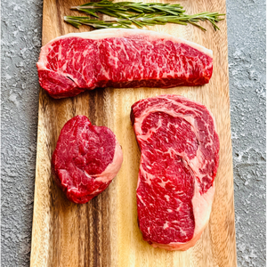 Three pieces of raw red meat on a wooden cutting board with rosemary, on a gray surface.