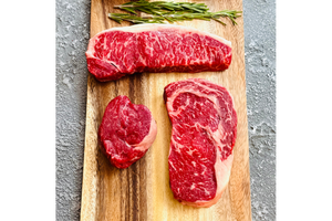 Three pieces of raw red meat on a wooden cutting board with rosemary, on a gray surface.